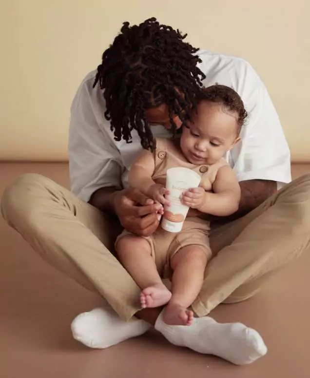 An adult and a baby sit on a beige floor. The adult, wearing a white shirt and beige pants, lovingly holds the child in their lap. The baby, dressed in a brown outfit, is holding a GRIN product tube and curiously looking at it.